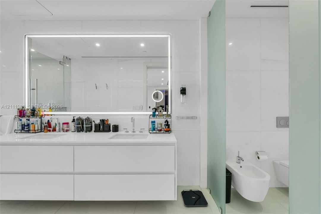 Floating white vanity with dual rectangular undermount sinks, large LED-backlit mirror, wall-to-wall stone-finish tiling, and wall-mounted bidet and toilet