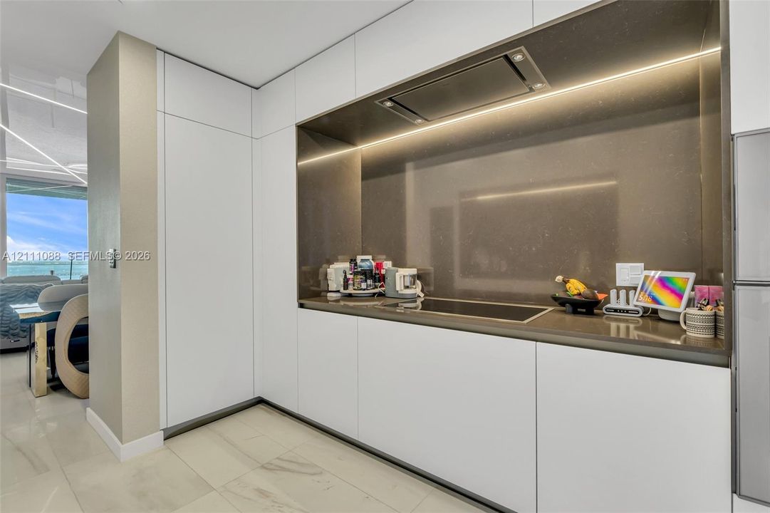 Sleek kitchen featuring matte white flat-panel cabinetry, a recessed cooktop, integrated range hood, solid-surface backsplash, and polished stone-finish flooring