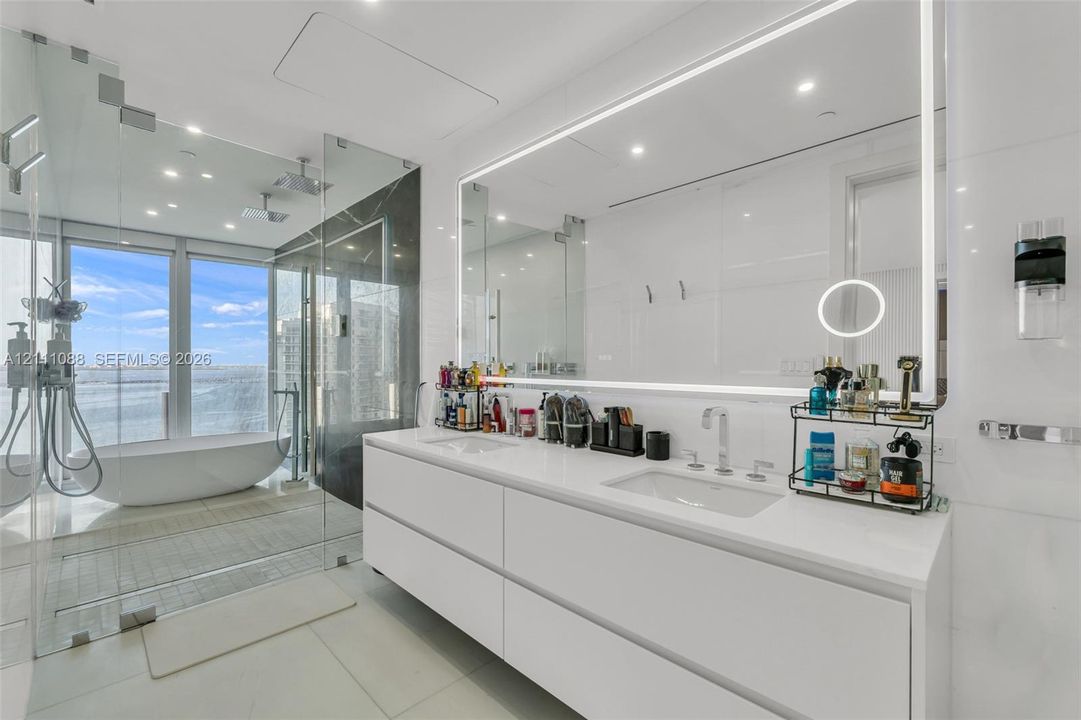 Floating double vanity with dual undermount sinks, expansive LED-backlit vanity mirror, glass-enclosed wet room featuring a freestanding soaking tub, and floor-to-ceiling windows