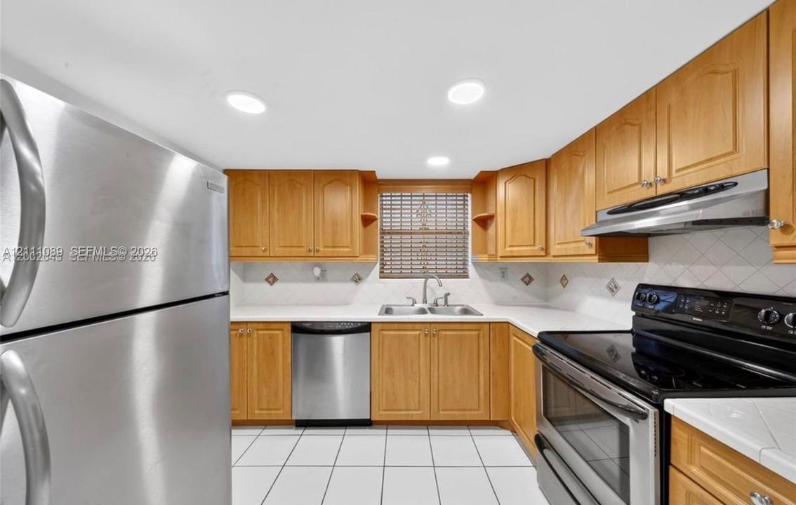 Kitchen featuring wood cabinetry, stainless steel appliances, white tile countertops with a diamond-patterned backsplash, a double-basin stainless steel sink, and tile flooring