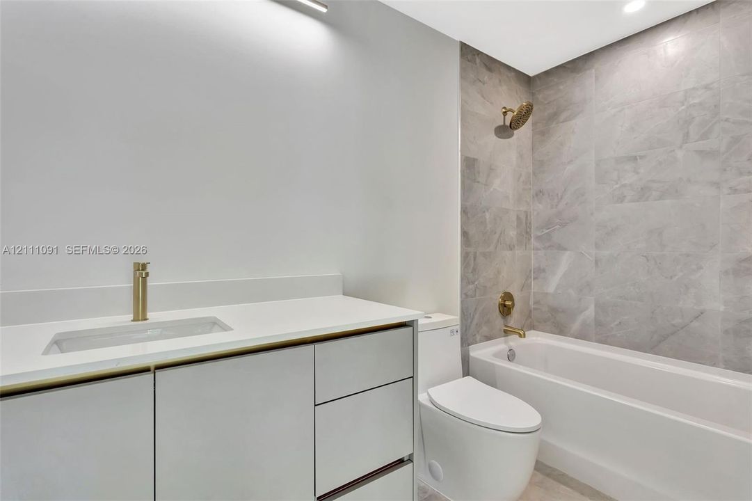 Bathroom featuring a vanity with quartz countertop, light-toned cabinetry, brass-finish fixtures, and a tub enclosure with large-format tile surround