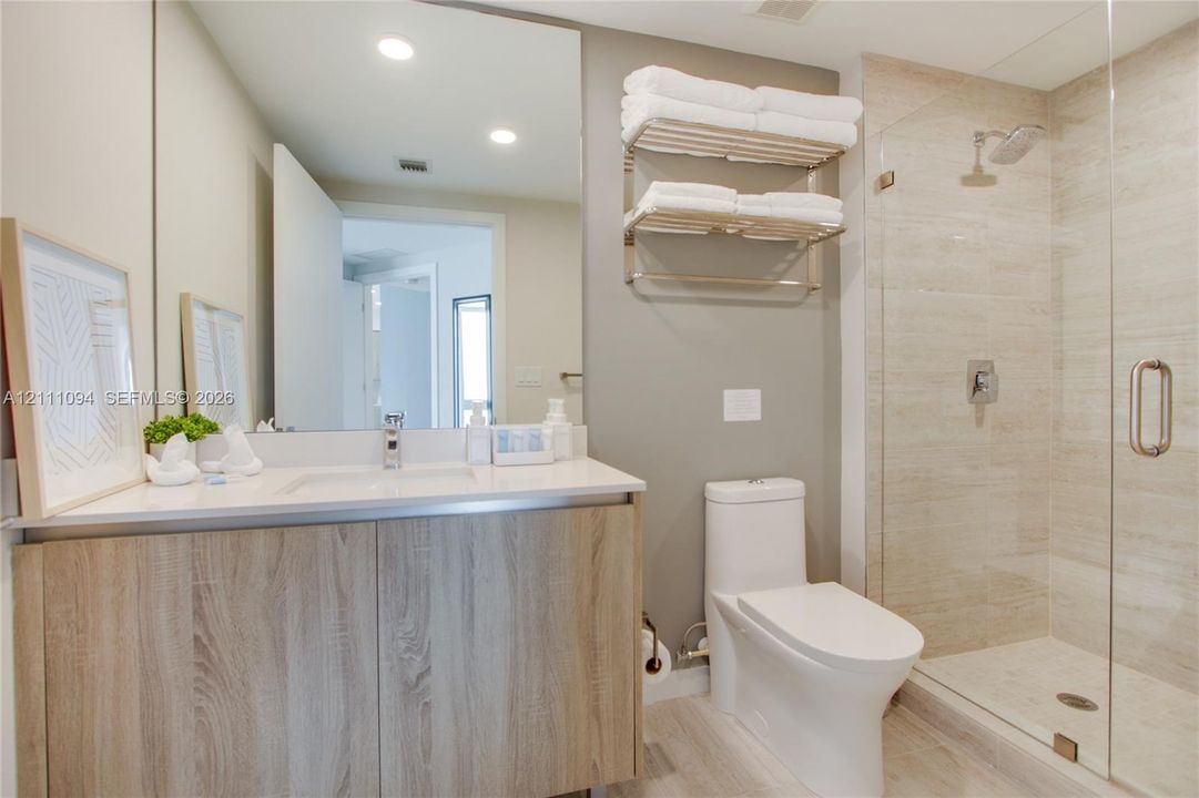 Bathroom featuring a wood-finish vanity, a solid surface countertop with an integrated sink, a large wall-mounted mirror, a walk-in shower with floor-to-ceiling tile, and a wall-mounted metal towel rack