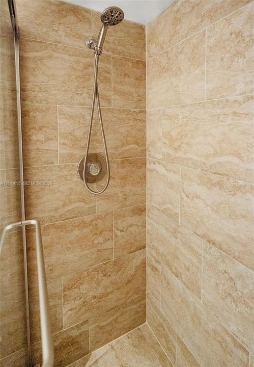Shower enclosure featuring floor-to-ceiling stone-finish tile, adjustable handheld shower fixture, circular polished metal control valve, and semi-frameless metal door hardware