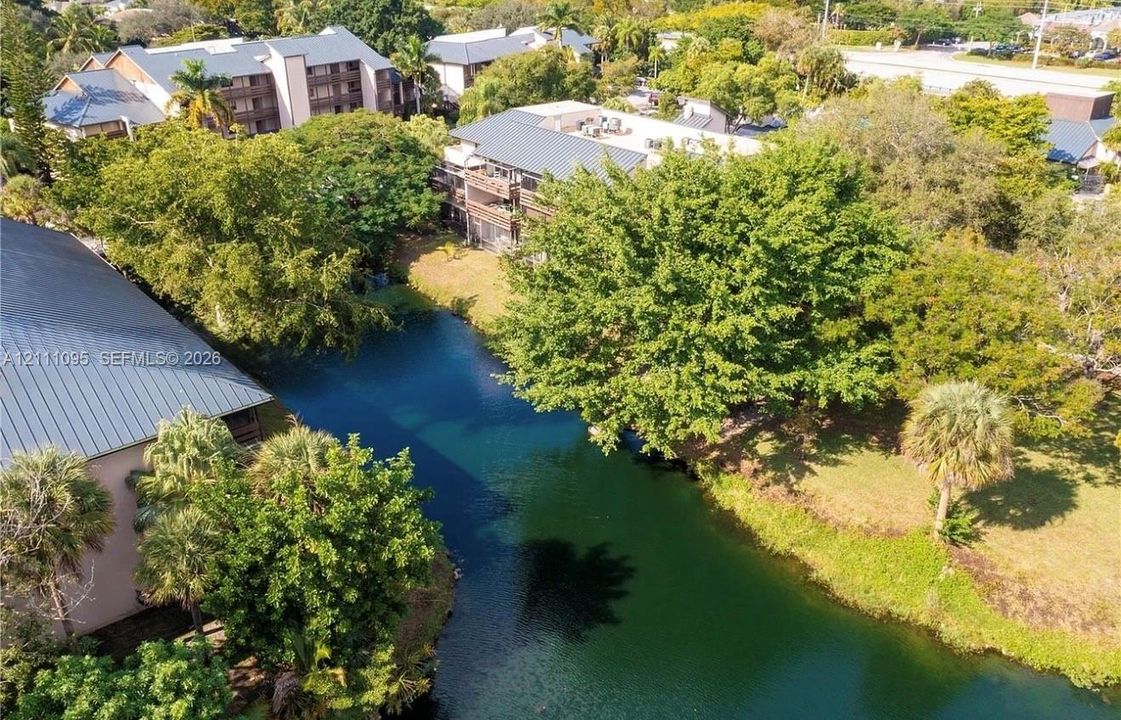 Aerial perspective featuring a multi-story building with a metal roof, situated alongside a water feature, accented by mature trees and palm foliage