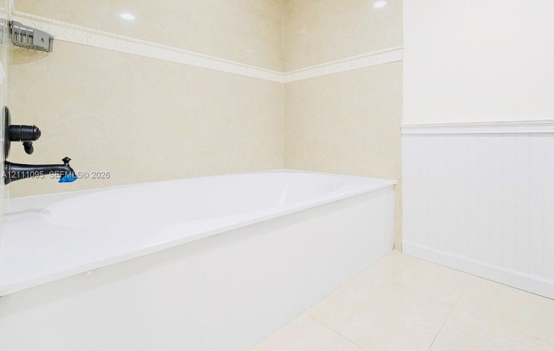 Built-in soaking tub with a wall-mounted matte black faucet, ceramic wall tile surround, integrated metal soap dish, and white wainscoting