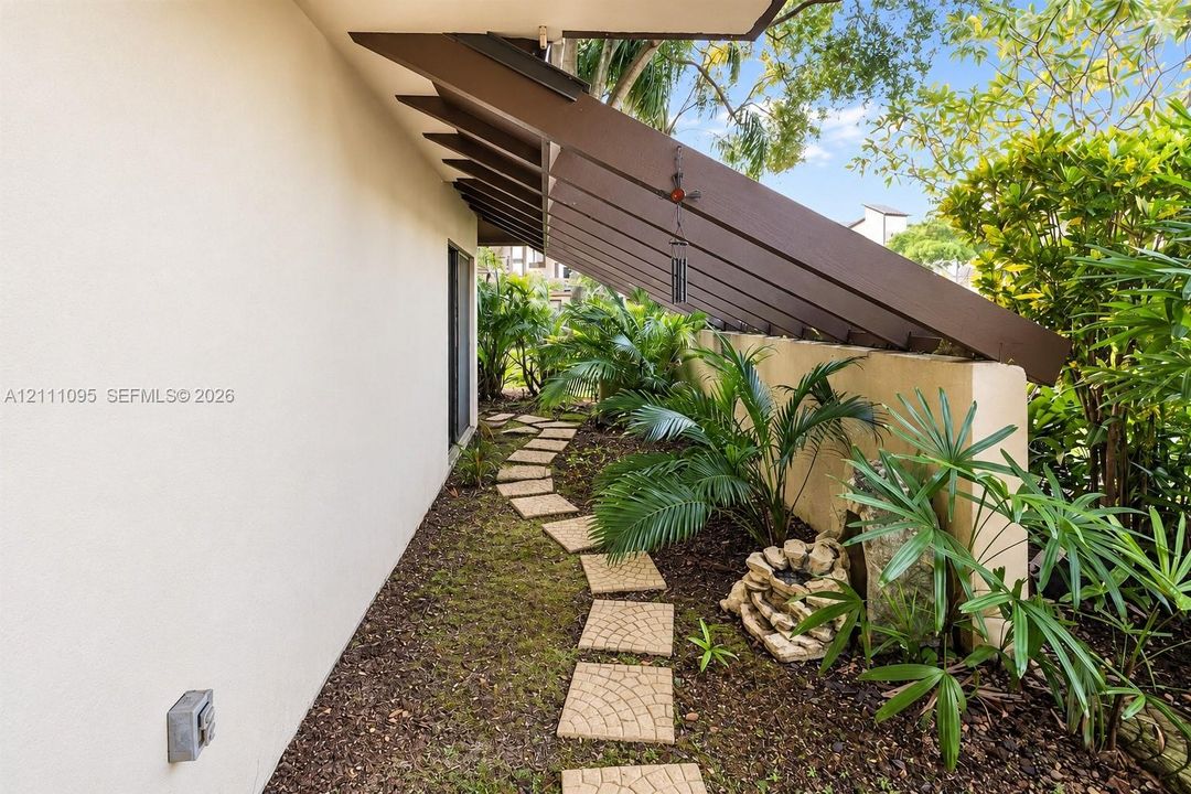 Exterior walkway featuring a stone paver path, exposed timber beams, stucco wall finish, and mature landscaping