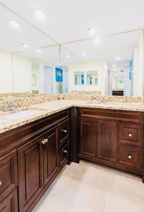 Double vanity featuring dark wood-finish cabinetry, stone-texture countertops with dual undermount sinks, and a large-format wall-to-wall mirror with integrated recessed lighting