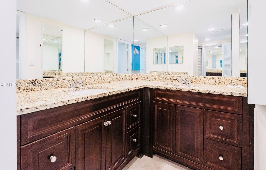 L-shaped vanity featuring dark wood-finish cabinetry, a stone-composite countertop, dual undermount sinks with metal hardware, and an expansive wall-to-wall mirror