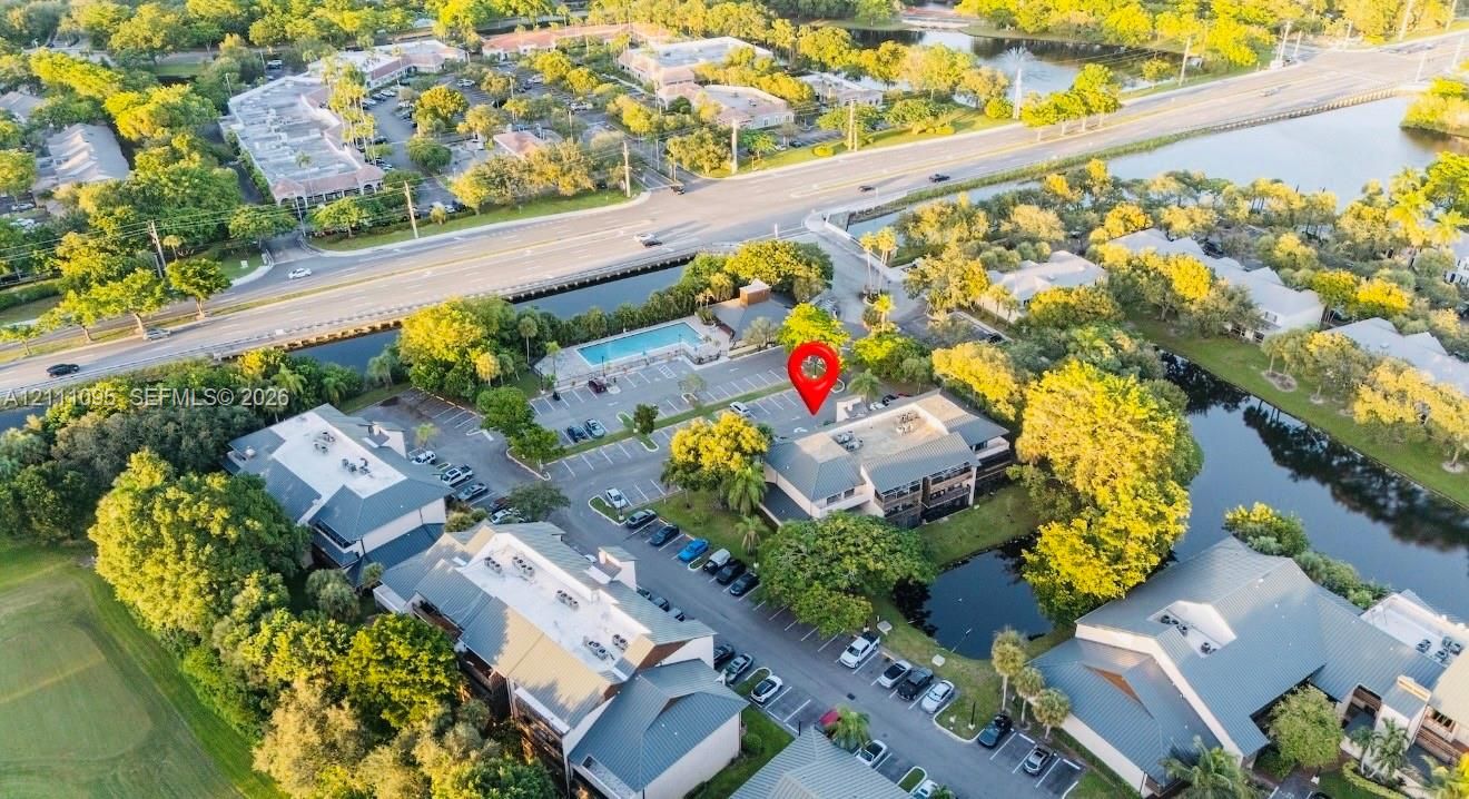 Aerial perspective featuring a multi-story building, an outdoor swimming pool, paved parking areas, and surrounding waterways