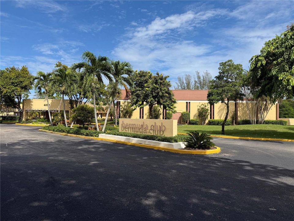 Entrance featuring paved drive, stone monument signage, manicured landscaping, and mature palm trees