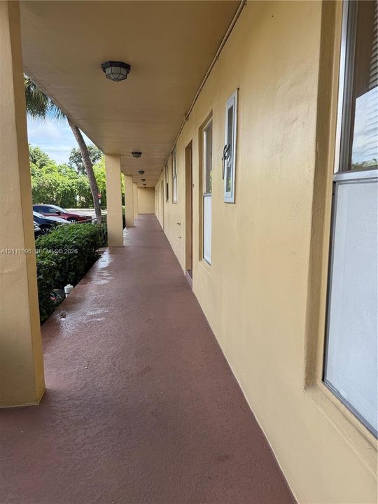 Covered exterior walkway featuring textured concrete flooring, flush-mount lighting, and solid structural pillars