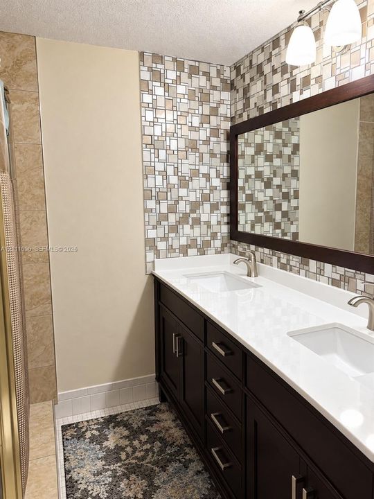 Double vanity featuring a white solid-surface countertop, dual rectangular undermount sinks, espresso-toned cabinetry with modern hardware, and a glass-tiled mosaic backsplash accented by a wood-framed wall mirror