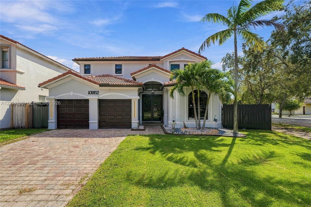 Two-story exterior featuring a tile roof, brick paver driveway, two-car garage with paneled doors, arched window detailing, and landscaped lawn with mature palm trees