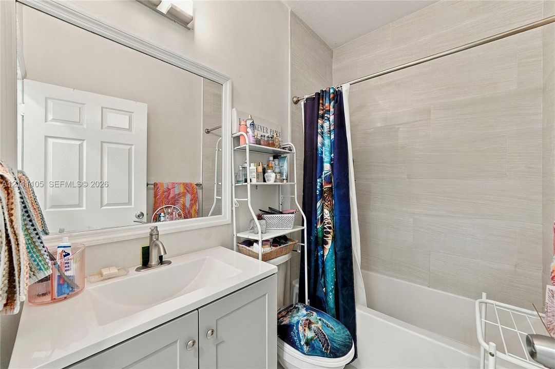 Shaker-style vanity with a solid-surface countertop, rectangular integrated basin, framed wall mirror, and neutral oversized horizontal wall tile surrounding the bathtub