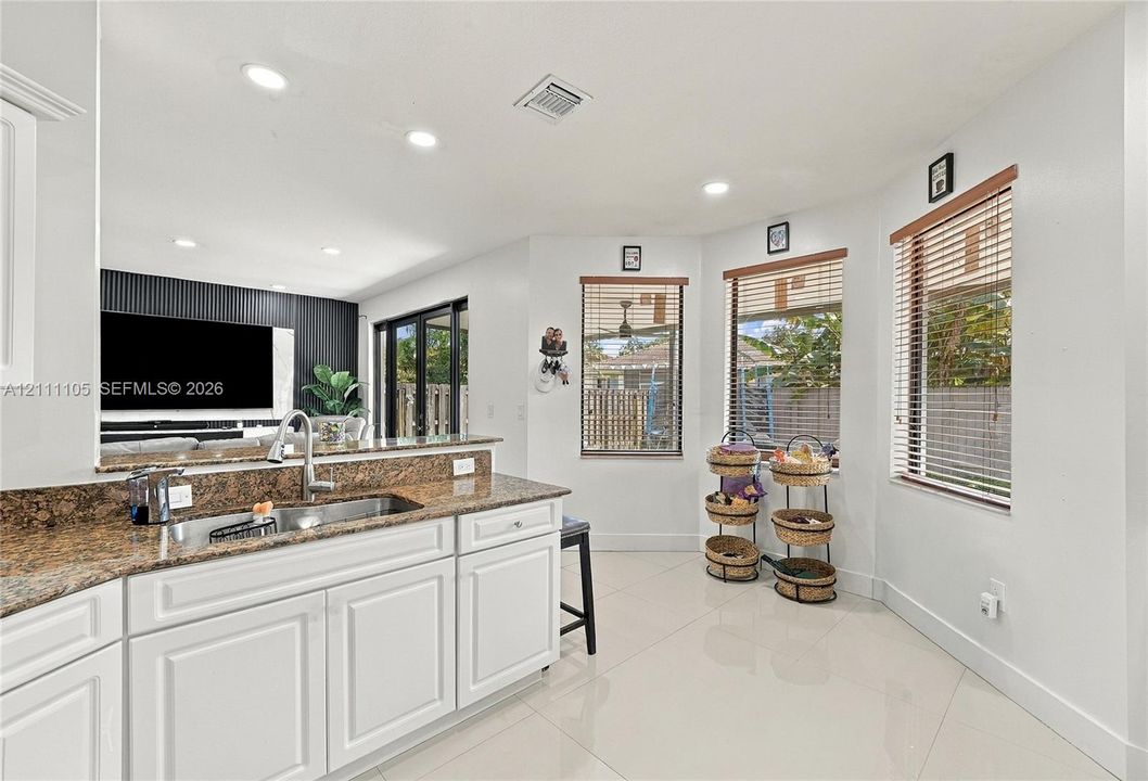 Kitchen featuring white raised-panel cabinetry, stone countertops, stainless steel undermount sink with goose-neck faucet, ceramic tile flooring, and multiple windows with horizontal blinds