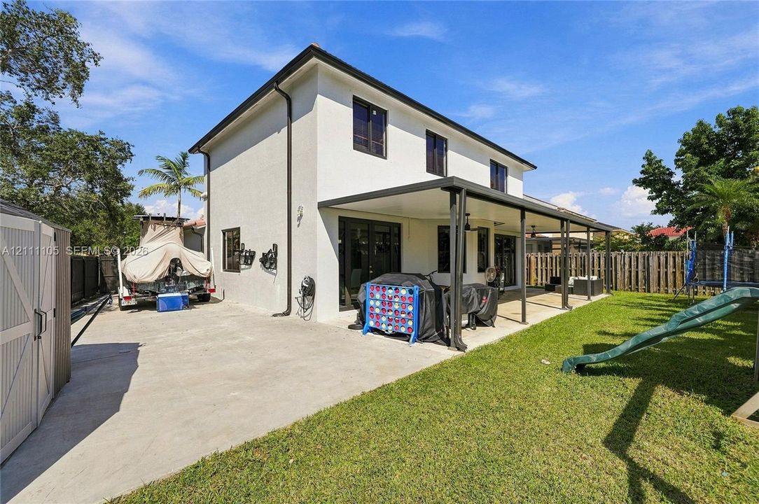 Two-story exterior featuring a light-toned stucco facade, a concrete patio, a covered lanai with structural posts, and a wood privacy fence