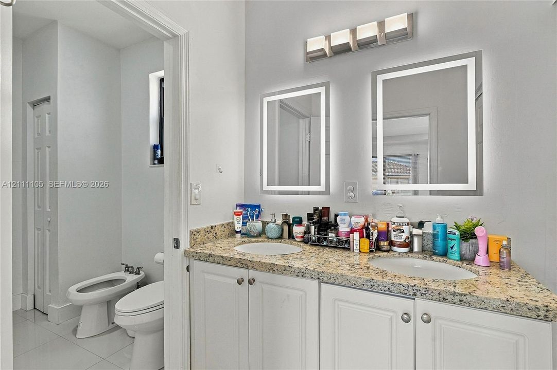 Dual-sink vanity with stone-finish countertop, white raised-panel cabinetry, two backlit rectangular mirrors, and four-light vanity fixture