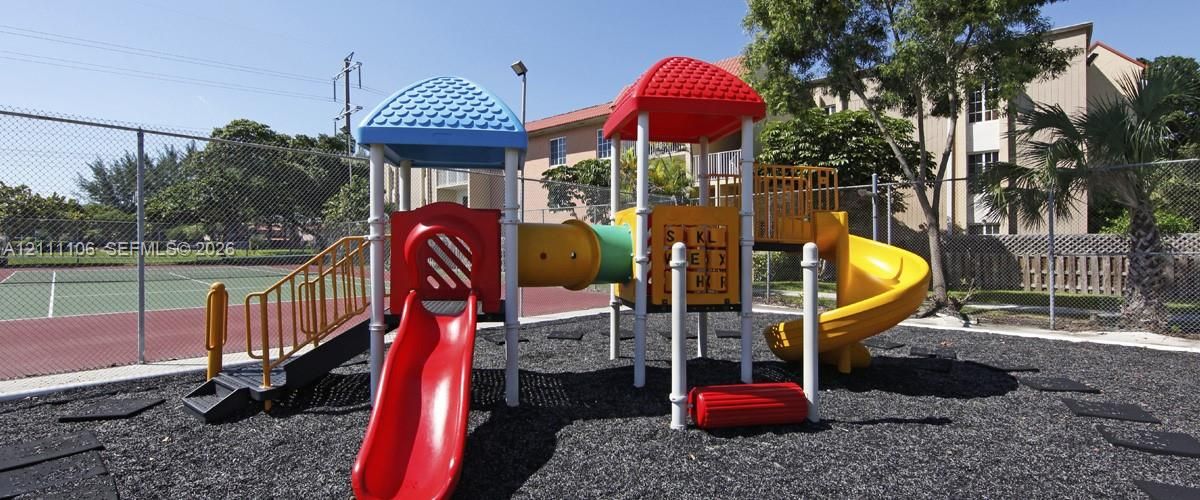 Outdoor recreation area featuring a multi-component play structure with slides and tunnels, rubberized surface ground cover, and chain-link fencing surrounding a hard-court surface