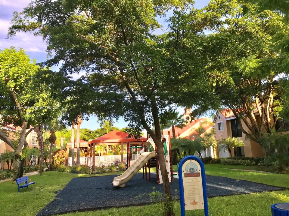 Outdoor recreation area featuring a playground structure with a slide, rubberized surfacing, covered gazebo, and mature canopy trees