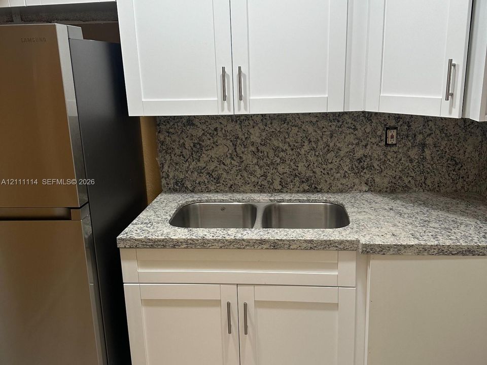 Shaker-style white cabinetry, stone-finish countertops, full-height stone backsplash, dual-basin stainless steel sink, and stainless steel refrigerator