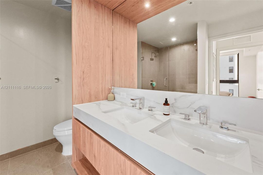 Double vanity featuring a light wood-finish cabinet, white solid-surface countertop with dual integrated sinks, polished chrome widespread faucets, and a full-width mirror