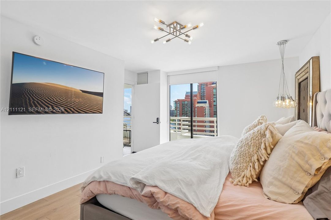 Light-filled room featuring wood-finish flooring, a multi-arm modern ceiling fixture, a suspended pendant light, and a sliding glass door leading to a private balcony