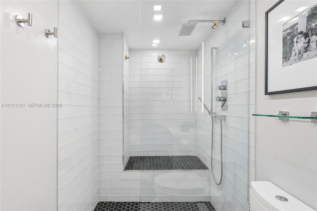 Walk-in shower featuring white subway wall tile, black hexagonal floor tile, glass enclosure, rainfall shower head, and hand-held shower fixture