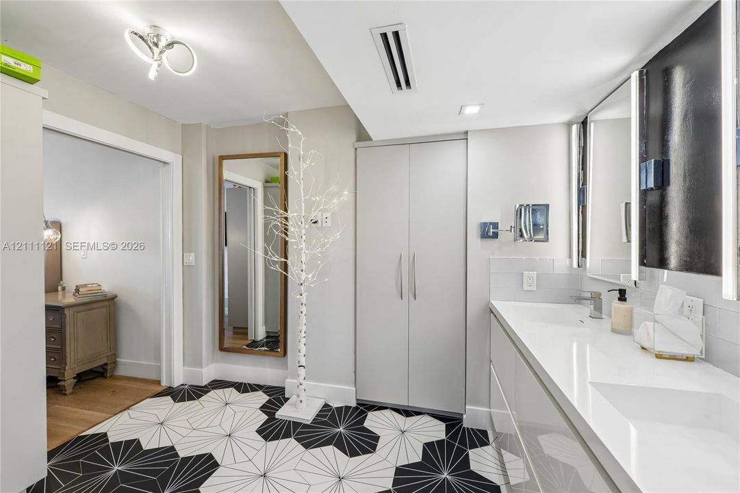 Bathroom featuring geometric patterned floor tile, integrated dual-sink vanity with a solid-surface countertop, full-width mirror with integrated lighting, and floor-to-ceiling built-in cabinetry
