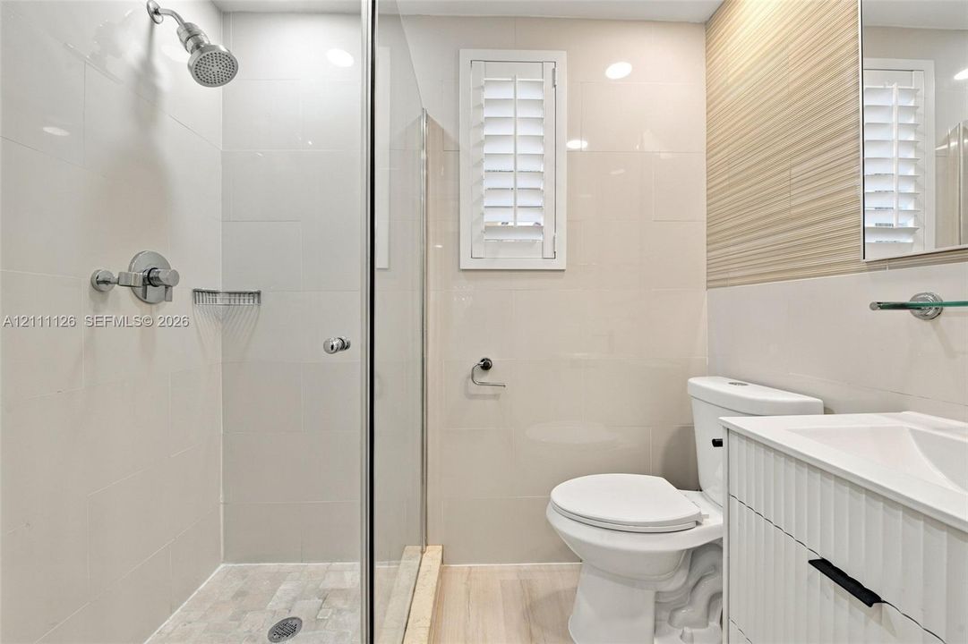 Walk-in shower with glass enclosure, wall-mounted shower head, textured accent wall, integrated vanity with white cabinetry, and shutter-style window treatment