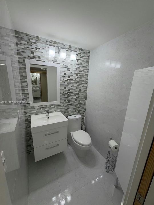 Modern vanity with a rectangular sink, multi-tone mosaic tile backsplash, polished light-colored floor tile, wall-mounted light fixture, and high-gloss cabinetry