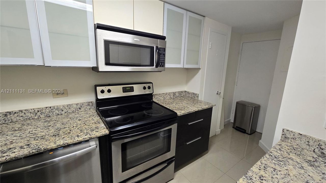 Kitchen featuring stone countertops, stainless steel appliances, upper cabinetry with glass-panel inserts, flat-panel lower cabinets, and tile flooring