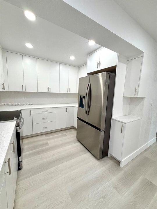 Kitchen featuring flat-panel white cabinetry, solid-surface countertops, recessed lighting, wood-finish flooring, and a stainless steel refrigerator