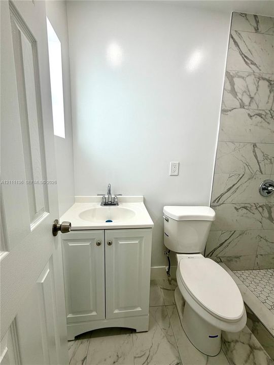 White vanity with a solid-surface countertop and integrated sink, stone-finish floor tiling, and a walk-in shower featuring large-format wall tiles and hexagonal floor tiling