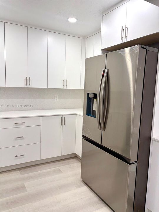 White cabinetry featuring linear hardware, light-toned solid surface countertops, light wood-finish flooring, and a stainless steel French door refrigerator with a built-in water dispenser