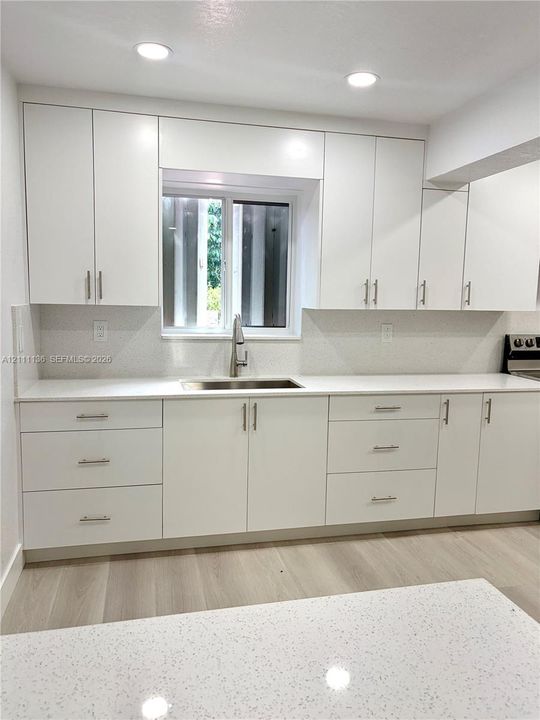 White flat-panel cabinetry featuring brushed nickel hardware, light-toned stone-finish countertops, stainless steel undermount sink with a pull-down faucet, and light wood-finish flooring