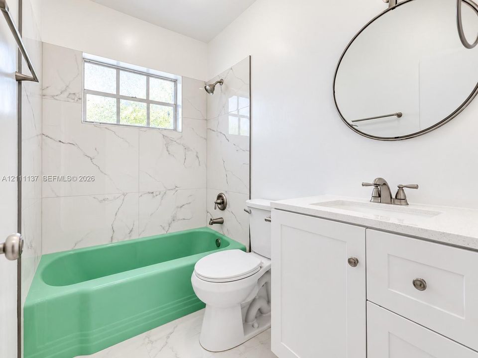 Bathroom featuring a white vanity with a composite countertop, oval wall-mounted mirror, large-format tile tub surround, and a colored bathtub