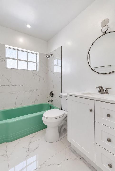 Bathroom featuring white tile flooring and wall surrounds with grey veining, a colored soaking tub, a white vanity cabinet with a stone-finish countertop, and a circular wall-mounted mirror