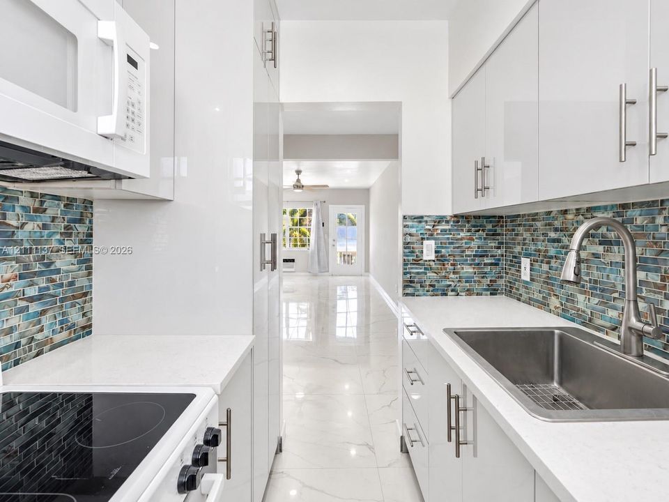 Kitchen featuring high-gloss white cabinetry, white quartz countertops, multi-tonal glass tile backsplash, stainless steel undermount sink with arched pull-down faucet, and polished stone-finish flooring