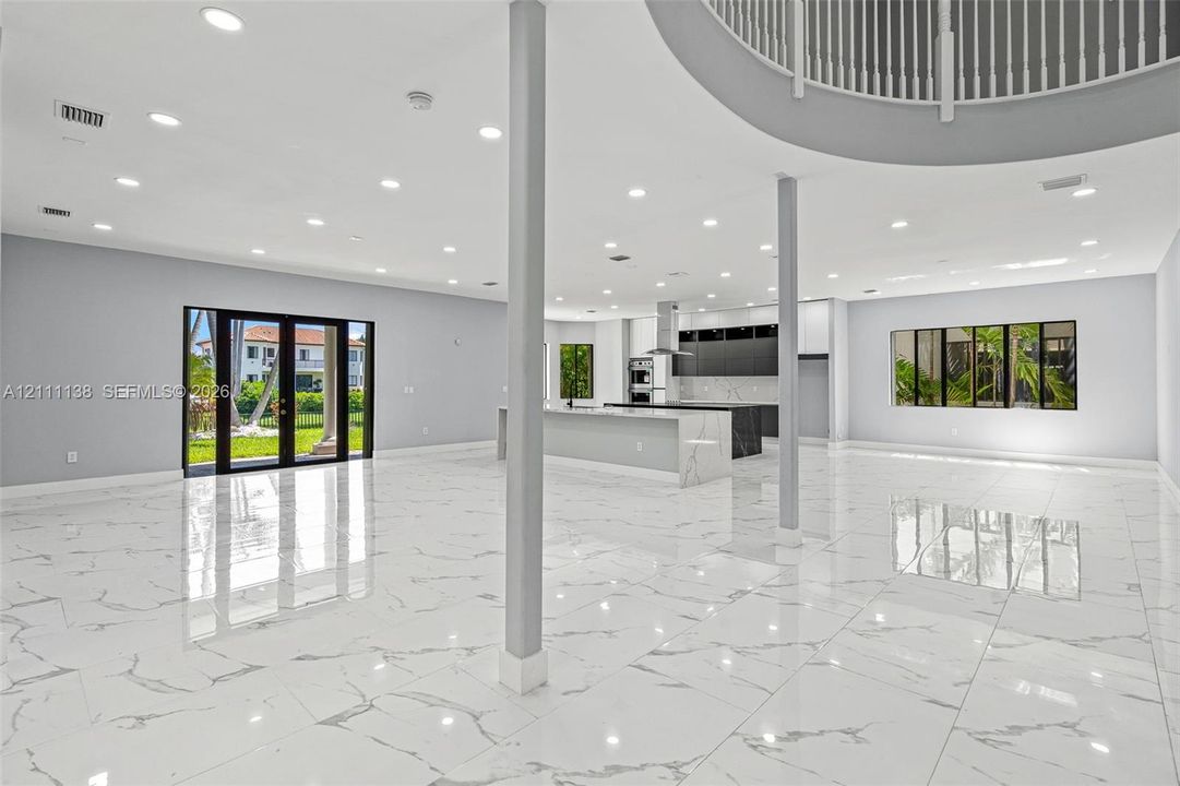 Open-concept interior featuring large-format stone-finish flooring, recessed lighting, a kitchen island with stone-finish countertop, structural support columns, and a mezzanine balcony with a metal railing