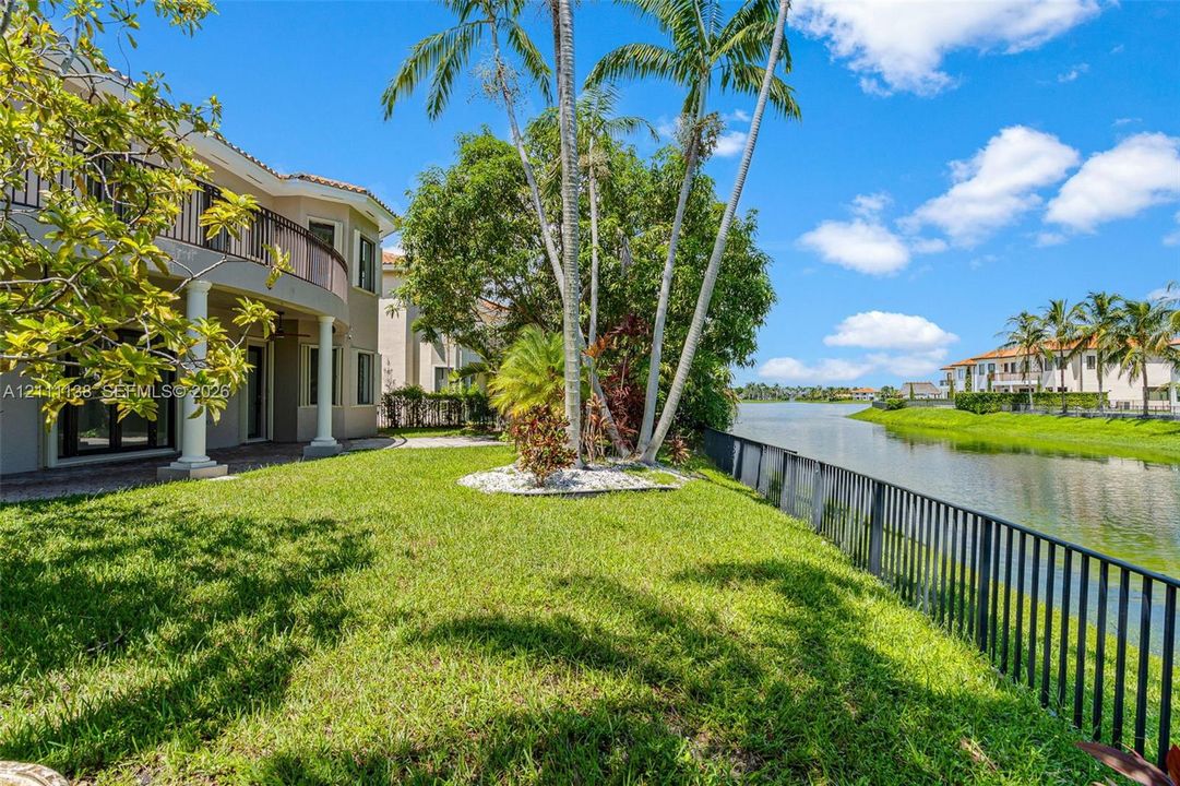 Waterfront backyard featuring a manicured lawn, metal perimeter fencing, mature palm trees, and a two-story exterior with a curved balcony