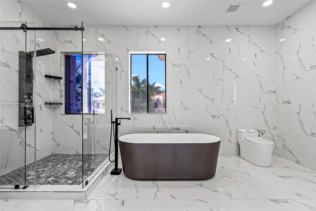 Full-height, large-format stone-patterned wall tile, freestanding soaking tub with floor-mounted matte black faucet, glass-enclosed walk-in shower with matte black rainfall showerhead, and hexagonal mosaic floor tile