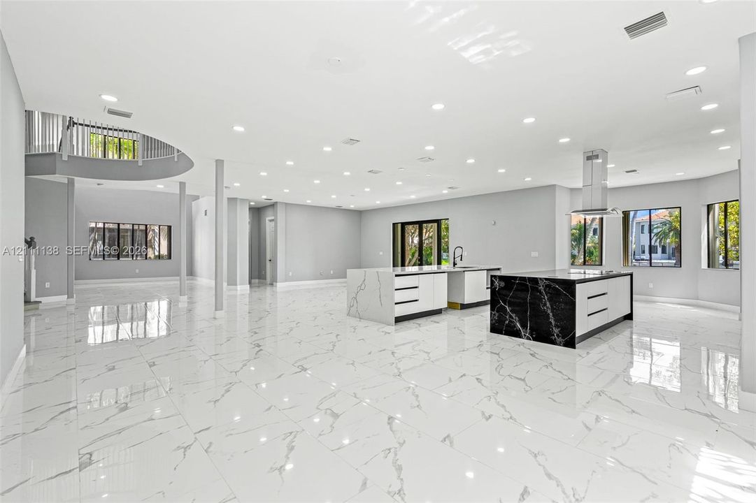Open-concept living space featuring large-format tile flooring, dual kitchen islands with contrasting waterfall-edge stone surfaces, recessed ceiling lighting, and a curved staircase with a metal balustrade