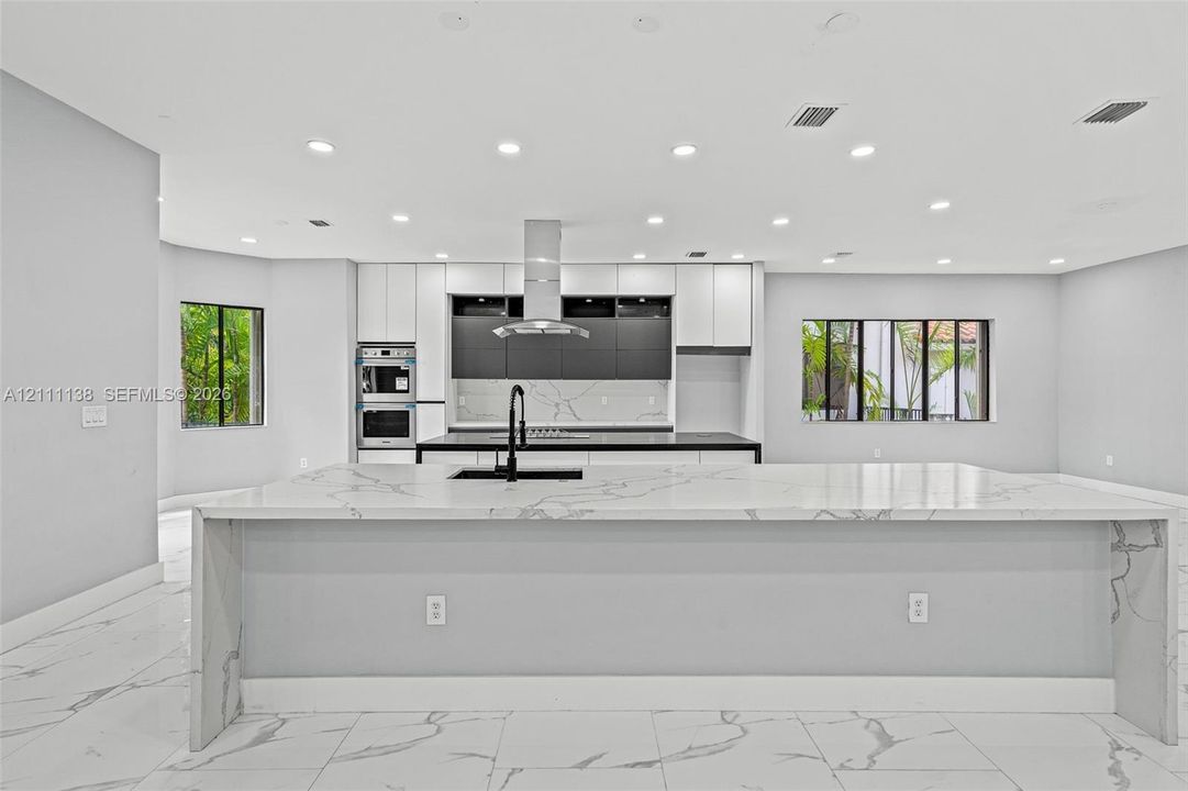 Kitchen featuring a large waterfall-edge island with stone-finish countertops, matte black faucet, integrated stainless steel double oven, two-tone cabinetry, and recessed lighting