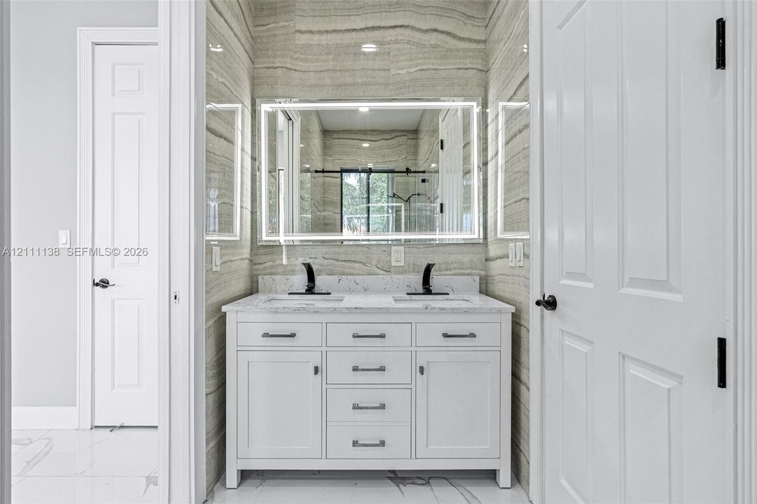 Double vanity featuring a white cabinetry base, stone-look countertop, integrated dual sinks, and matte black widespread faucets, accented by full-height linear pattern tile walls and a modern LED-backlit mirror