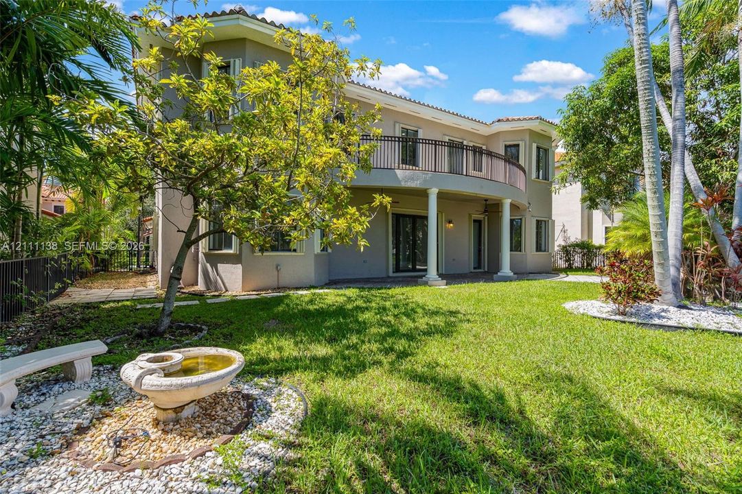Two-story stucco exterior featuring a curved upper-level balcony with metal railing, classic columns, stone-accented landscaping, and a tiered fountain
