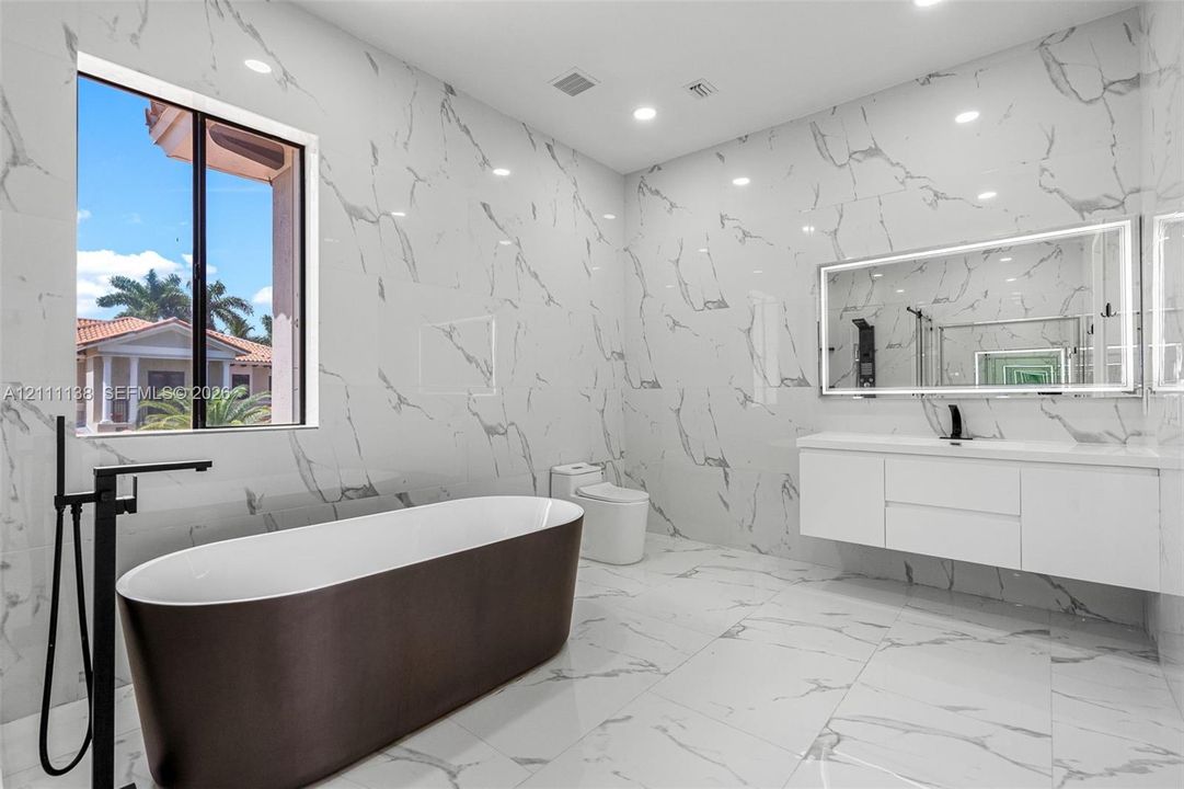 Bathroom featuring floor-to-ceiling large-format tile with stone-pattern finish, wall-mounted floating vanity, freestanding soaking tub with matte black floor-mounted faucet, and an oversized LED-backlit mirror