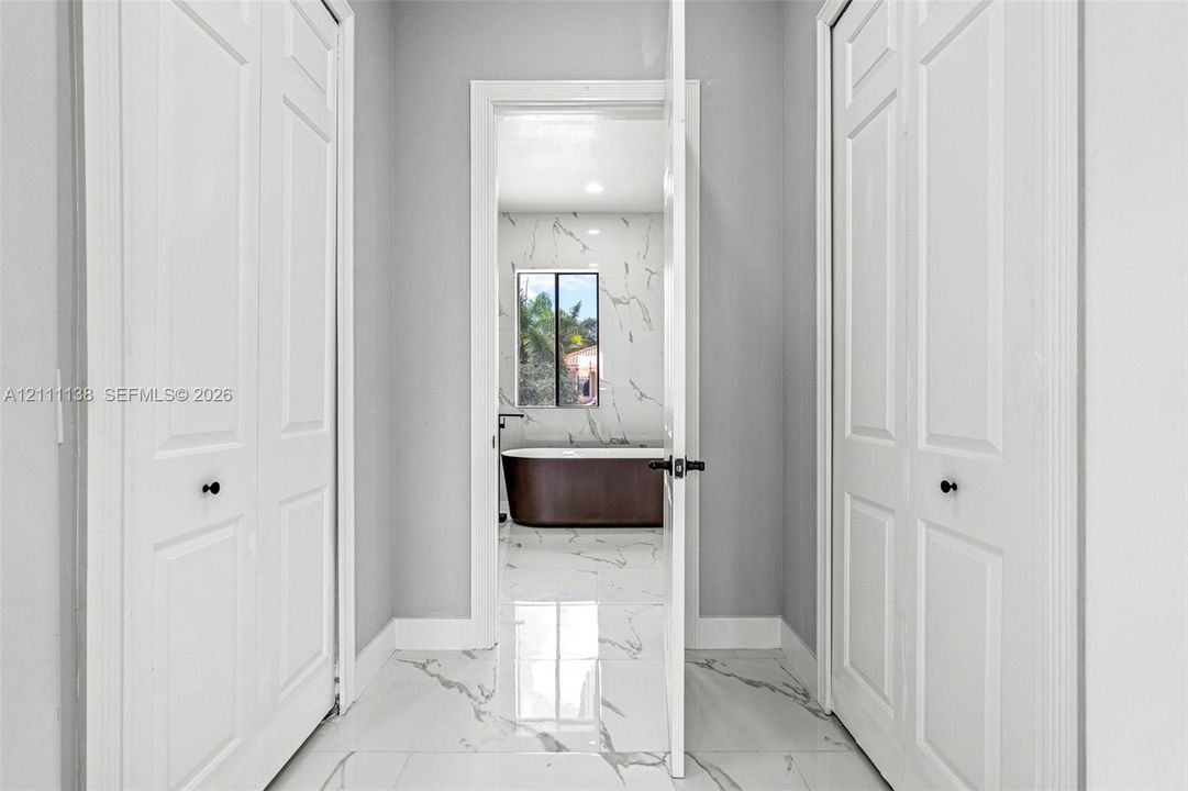 Hallway featuring white panel doors with matte black hardware and polished stone-finish floor tile with veining, leading into a space with matching stone-finish wall tile and a freestanding soaking tub