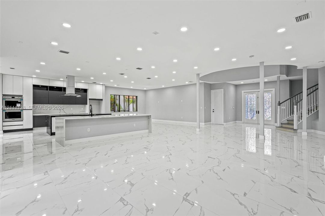 Open-concept interior featuring large-format tile flooring, recessed lighting, a modern kitchen with a dual-tone cabinetry finish, a central island, and a staircase with a wooden handrail