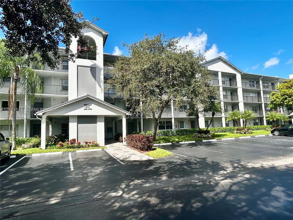 Multi-story residential building exterior featuring a covered entryway, private balconies, paved parking area, and mature landscaping