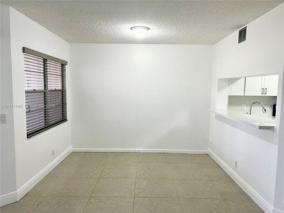 Neutral-toned floor tiling, window with slatted blinds, kitchen pass-through, and neutral-toned wall paint